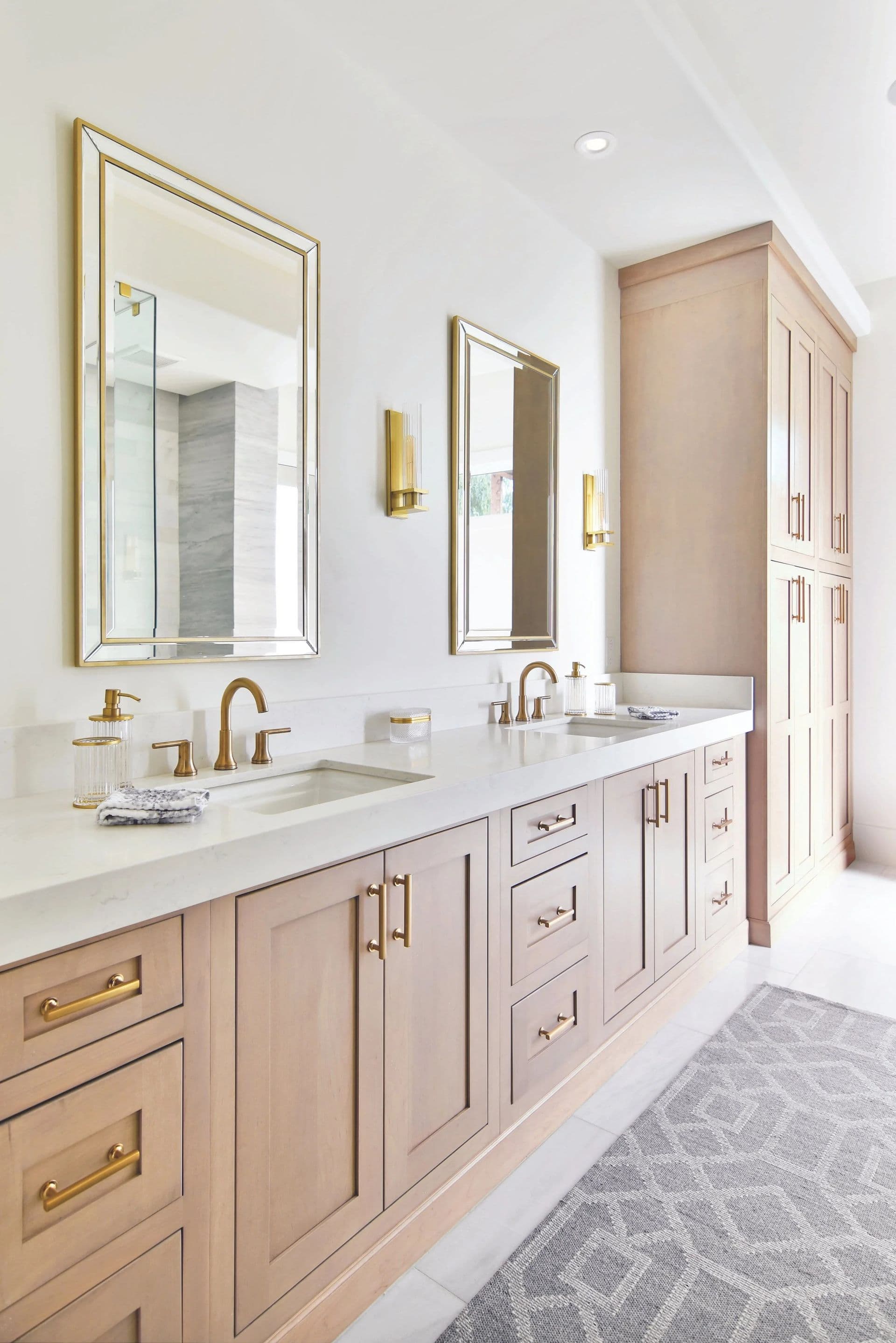 A newly-remodeled bathroom with light brown natural-colored cabinets, white countertops, double vanity, and gold fixtures.