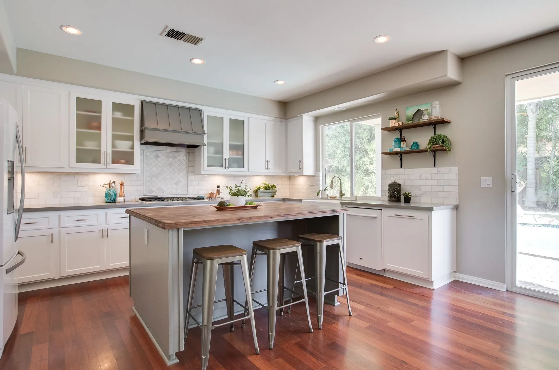 A kitchen with newly-refaced white cabinets, white marbled tile backsplash, central island with bar seating and wood countertop, and hardwood floor.