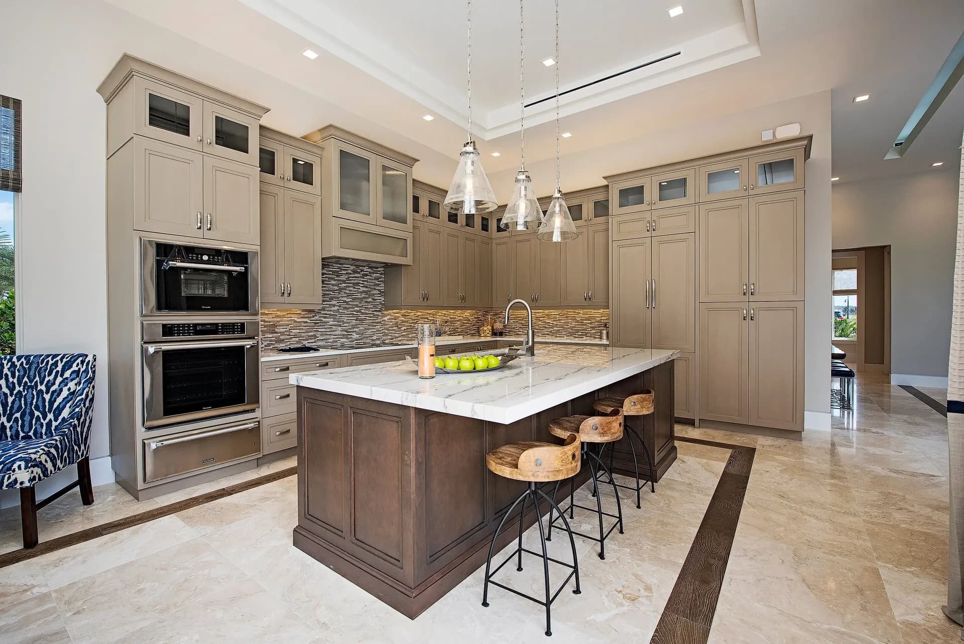 A light gray kitchen with beige cabinets, brown mosaic tile backsplash, central island with white marbled countertop and bar stool seating.